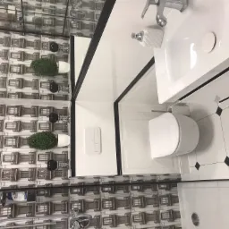 Nowoczesna łazienka z białą ceramiką, wzorzystą tapetą imitującą budynki, czarnymi akcentami i rośliną w doniczce.