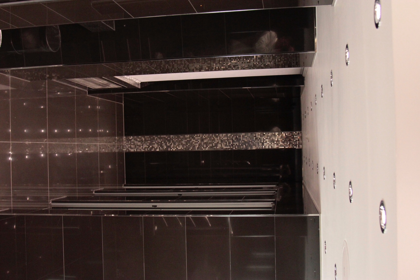 Ciemnobrązowa, prawie czarna, łazienka z błyszczącymi, prostokątnymi płytkami, mozaiką i oświetleniem punktowym w suficie