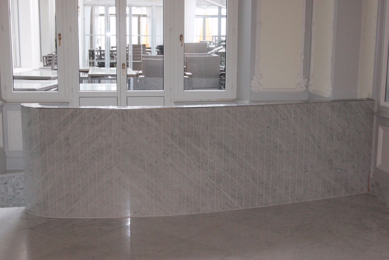 Zakrzywiona ściana wyłożona wąskimi, pionowymi płytkami marmurowymi w odcieniach szarości, z widocznymi drzwiami balkonowymi w tle.