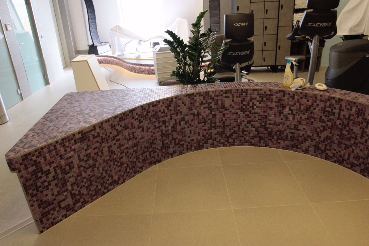 Zaokrąglona ściana wyłożona mozaiką w odcieniach fioletu i brązu, z beżową podłogą z płytek, w tle widoczne wyposażenie siłowni i elementy dekoracyjne.