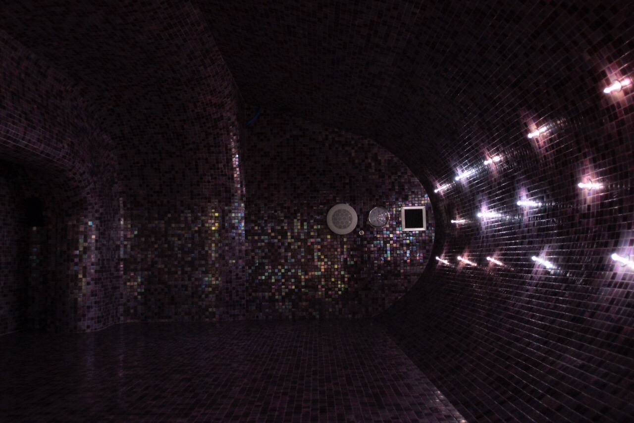 Ciemna, zaokrąglona przestrzeń wyłożona mozaiką z błyszczących płytek, oświetlona rzędem punktowych świateł wzdłuż jednej ze ścian, z widocznymi elementami wyposażenia na ścianie.