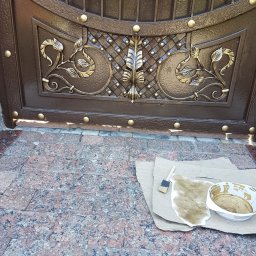 PERFEKT Sp. z o.o. - Fragment metalowej, brązowej bramy z dekoracyjnymi, złotymi elementami, z widocznymi śladami farby i narzędziami malarskimi na podłożu z kostki brukowej.