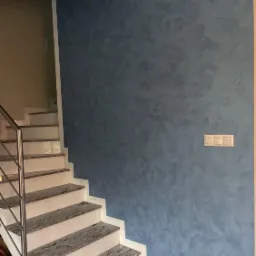 Ściana pomalowana dekoracyjną farbą w odcieniach niebieskiego, widoczne schody z szarymi stopniami z granitu i metalową balustradą, oświetlenie punktowe na ścianie.