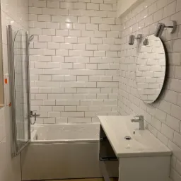 Nowoczesna łazienka w bieli z wanną, umywalką i lustrem owalnym, wykończona białymi, prostokątnymi płytkami ułożonymi w cegiełkę.