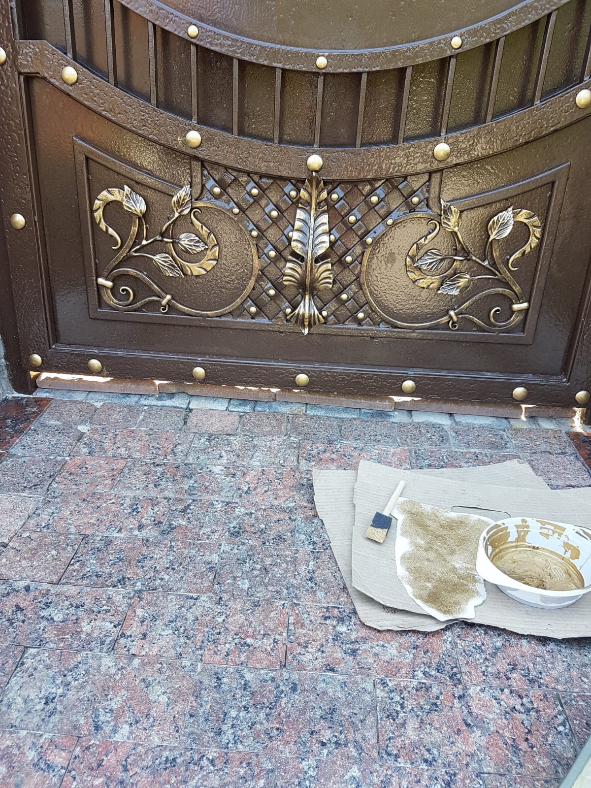 Fragment metalowej, brązowej bramy z dekoracyjnymi, złotymi elementami, z widocznymi śladami farby i narzędziami malarskimi na podłożu z kostki brukowej.