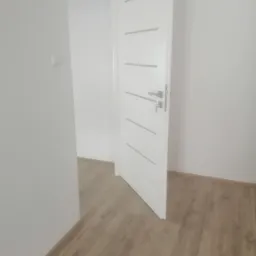 Otwarta biała, nowoczesna drzwi wewnętrzne z poziomymi frezami i srebrną klamką w nowym, jasnym pomieszczeniu z panelami podłogowymi.