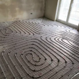 Rozłożone rury ogrzewania podłogowego na folii z siatką, przed wylaniem posadzki, widok z góry, surowe ściany w tle.