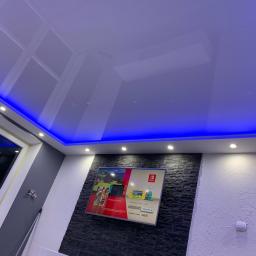 DANLUKS Usługi remontowo wykończeniowe - Nowoczesne wnętrze pokoju z podwieszanym sufitem z niebieskim oświetleniem LED, ściana z dekoracyjną okładziną kamienną pod telewizorem.