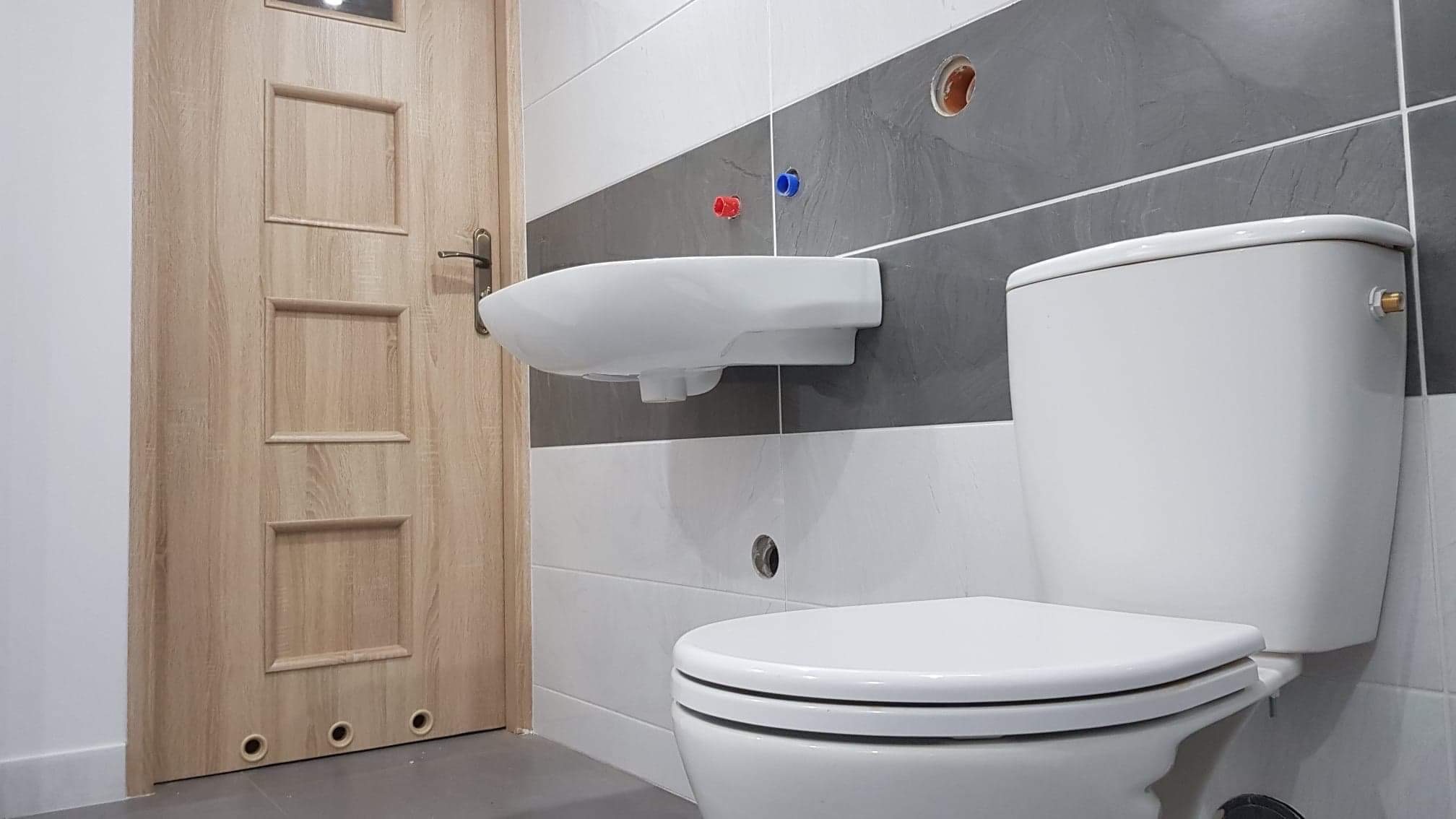 Nowoczesna łazienka w trakcie wykańczania: biała umywalka i toaleta na tle szaro-białych płytek, widoczne otwory na rury i drewniane drzwi z trzema okrągłymi otworami u dołu.