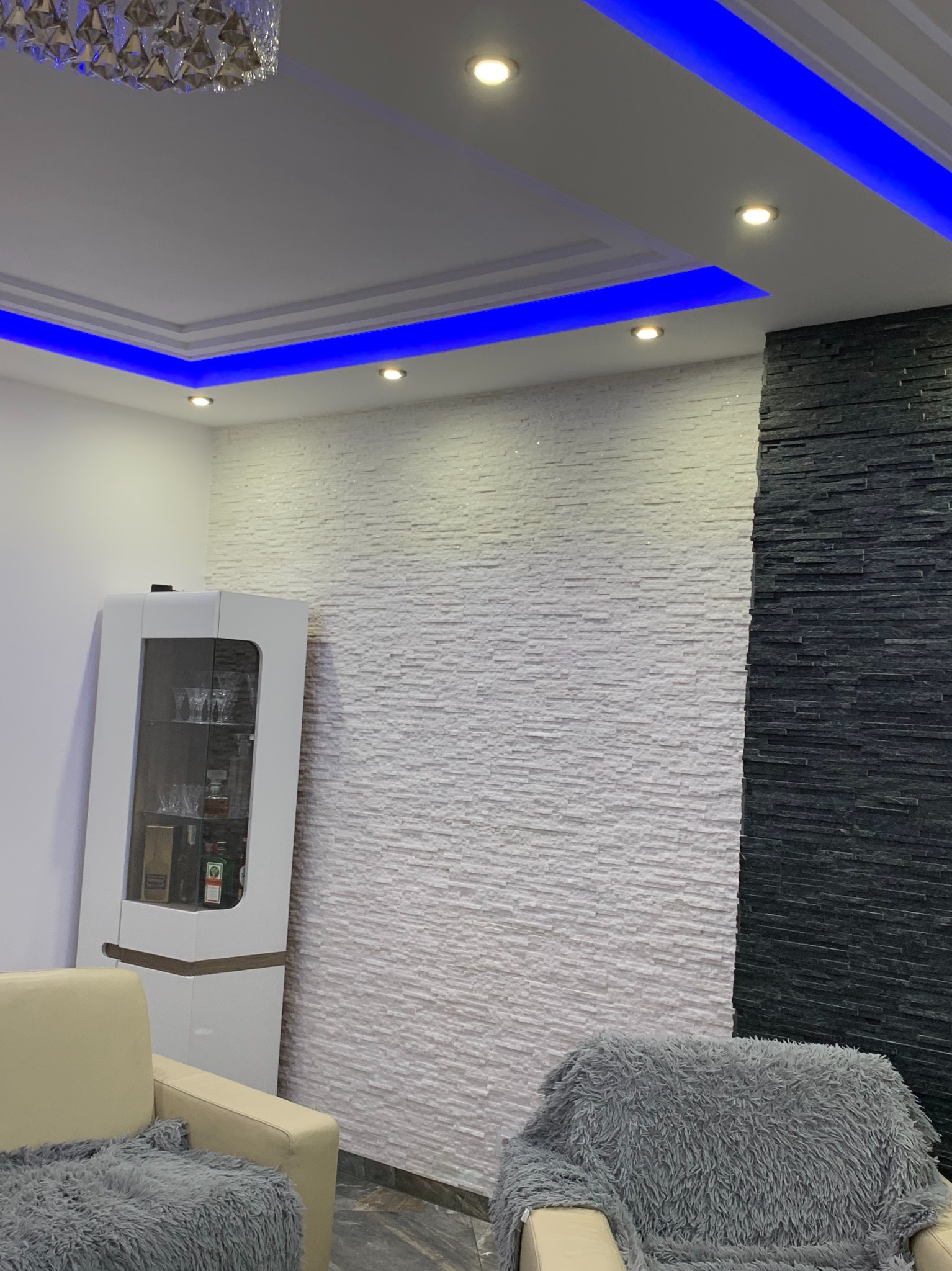 Nowoczesny salon z podświetleniem LED na suficie, ściana wykończona białym kamieniem dekoracyjnym i fragmentem czarnego kamienia łupkowego, obok biała witryna z napojami i fragment kremowej sofy...