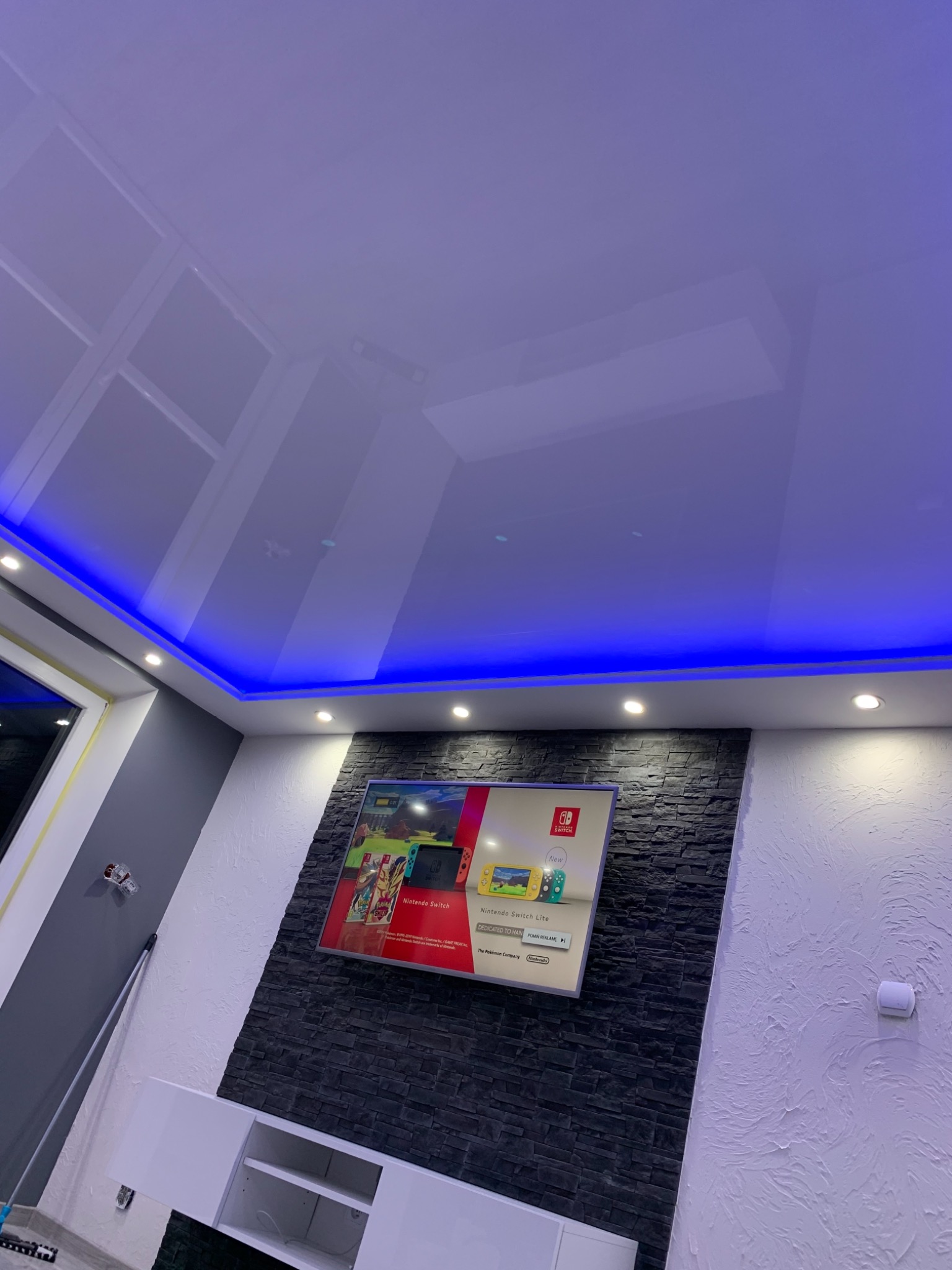 Nowoczesne wnętrze pokoju z podwieszanym sufitem z niebieskim oświetleniem LED, ściana z dekoracyjną okładziną kamienną pod telewizorem.