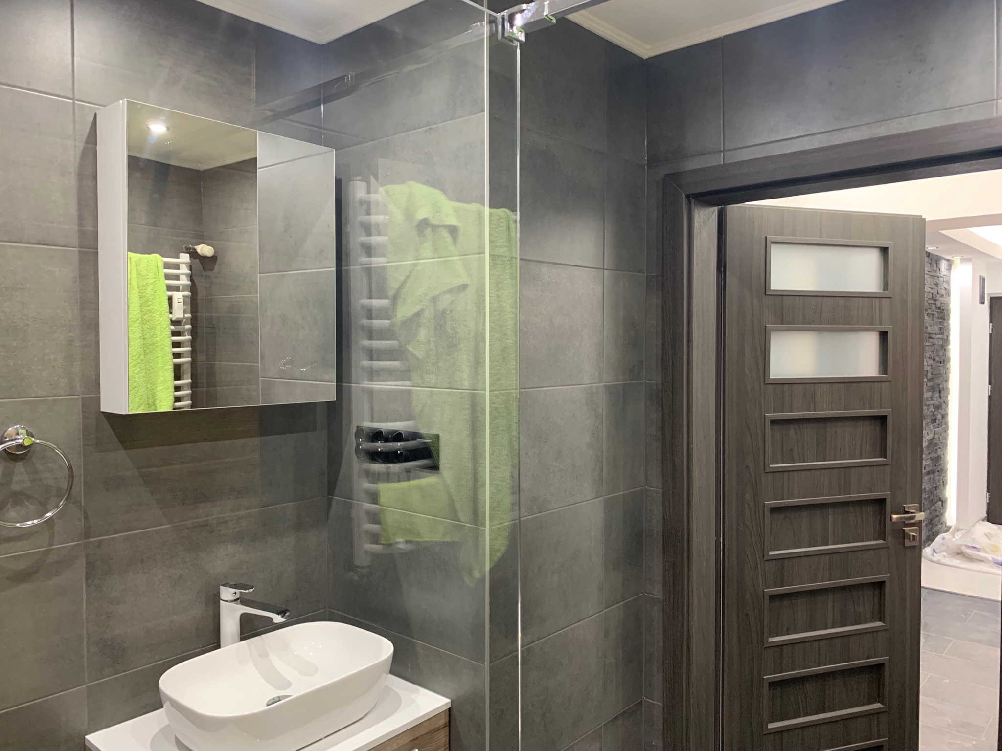 Nowoczesna łazienka z szarymi płytkami, umywalką nablatową, szklaną kabiną prysznicową i drewnianymi drzwiami z przeszkleniami.