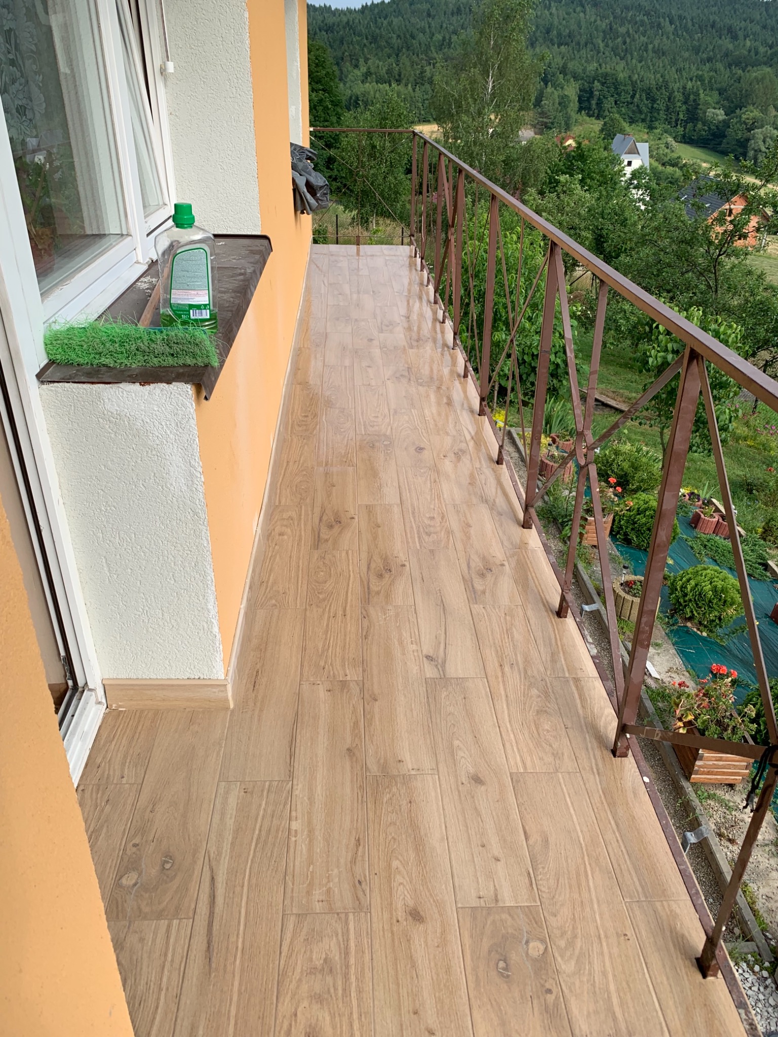 Błyszczące płytki imitujące drewno na wąskim balkonie z metalową balustradą, widok na zieleń i las w tle, butelka płynu i szczotka na parapecie.