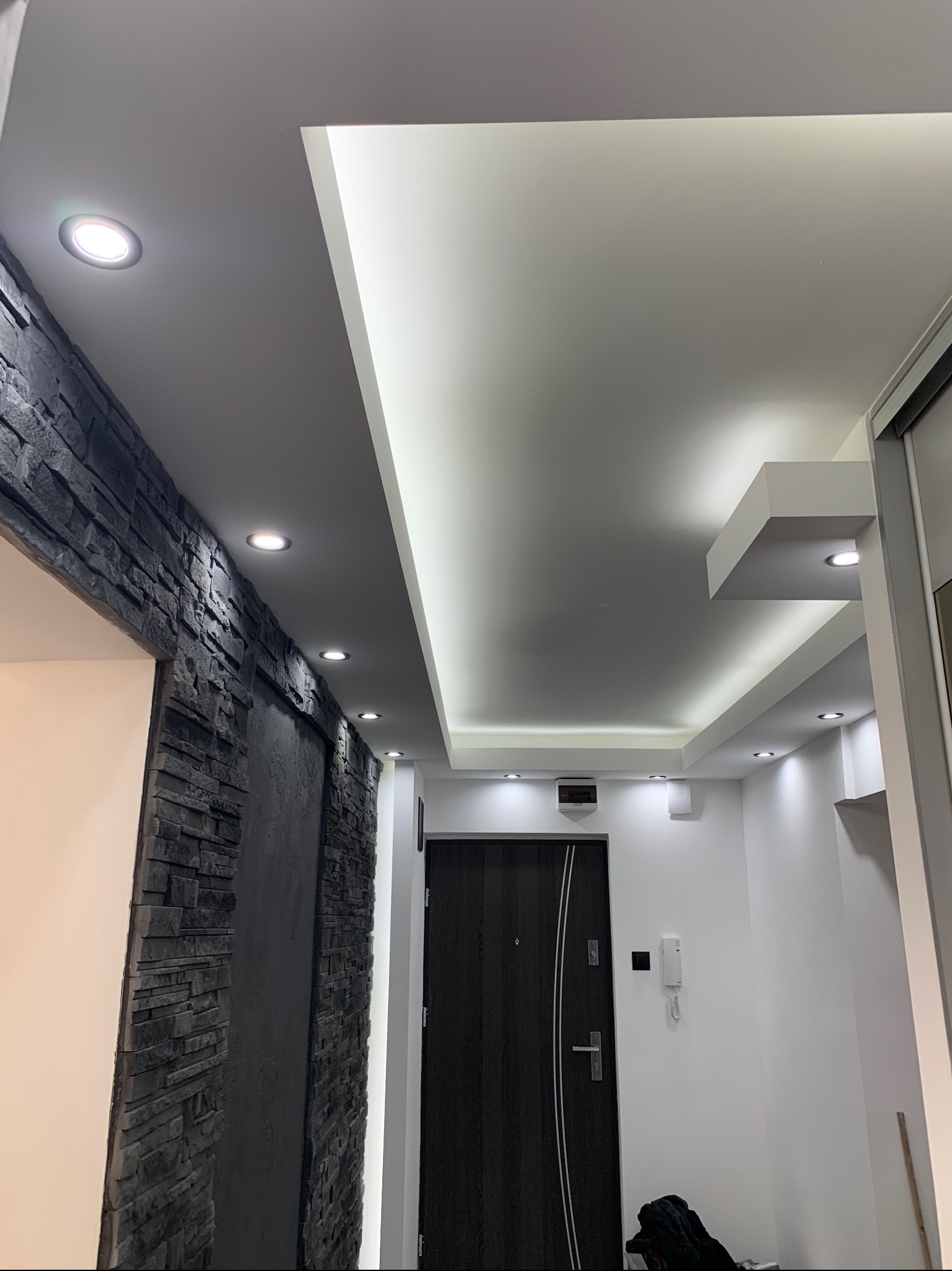 Nowoczesny przedpokój z podwieszanym sufitem z oświetleniem LED, ściana wykończona czarnym kamieniem dekoracyjnym, ciemne drzwi wejściowe z metalowymi akcentami.