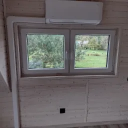 Biały klimatyzator zamontowany nad oknem w pokoju wykończonym jasnym drewnem, z widokiem na zielony ogród.