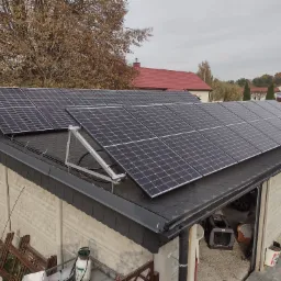 Panele słoneczne zamontowane na dachu garażu z bitumiczną dachówką, widoczne elementy konstrukcji mocującej, w tle drzewa jesienią i zabudowa jednorodzinna.