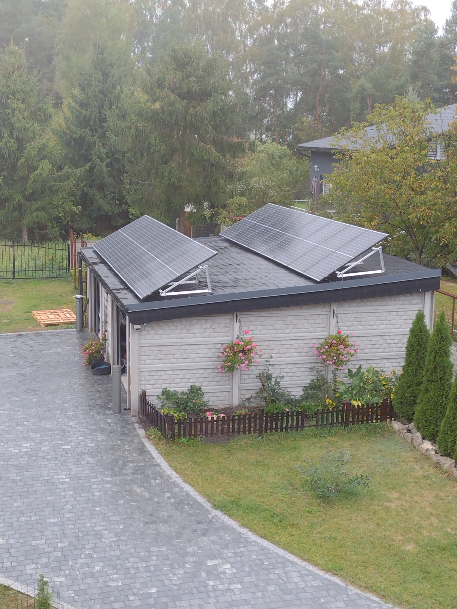 Dwa panele słoneczne zamontowane na dachu garażu z szarej cegły, widok z góry na posesję z kostką brukową i zielenią w pochmurny dzień.