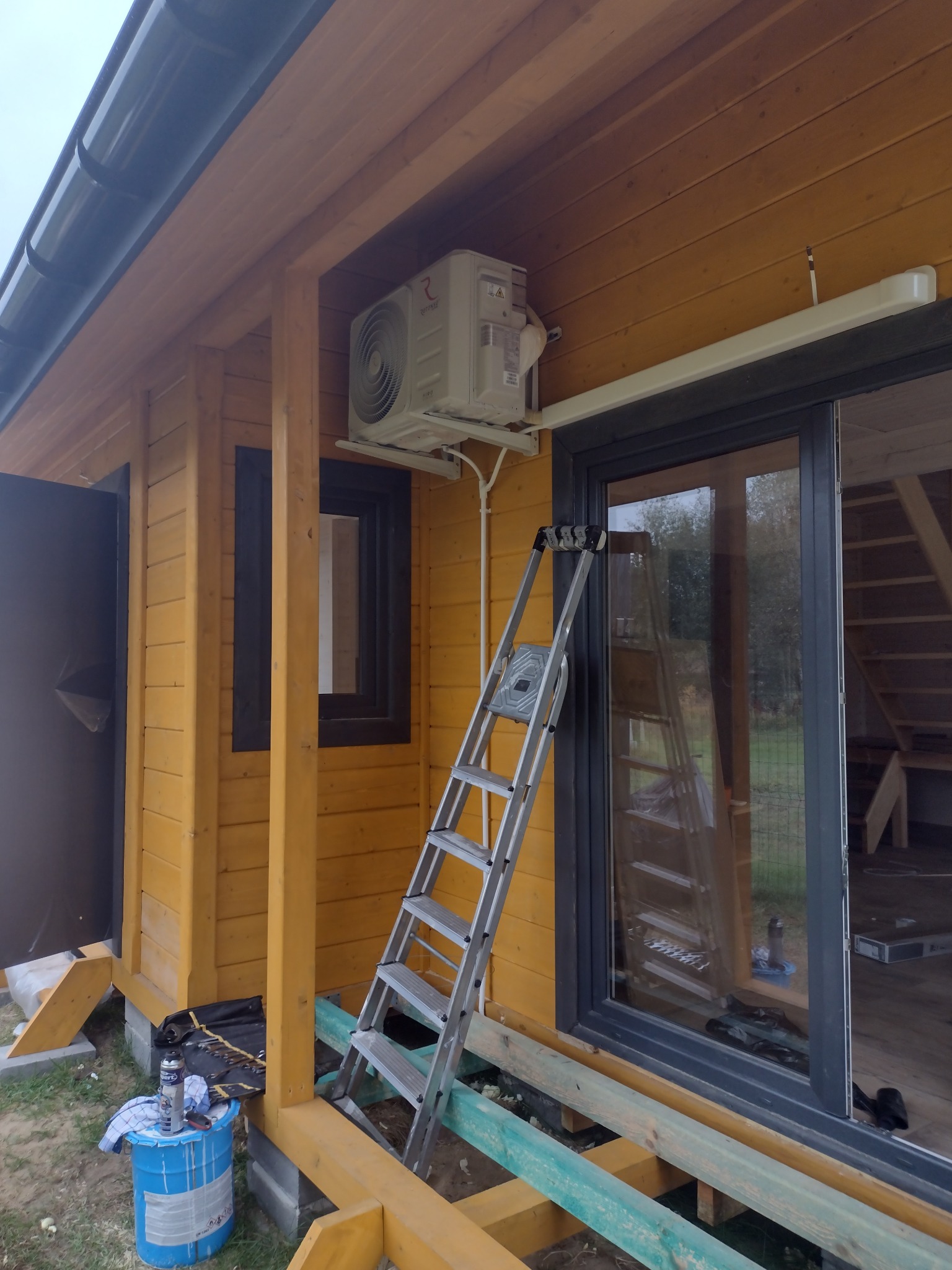 Montaż jednostki zewnętrznej systemu chłodzenia na drewnianej elewacji domu, widoczna drabina oparta o ścianę i narzędzia w niebieskim wiadrze