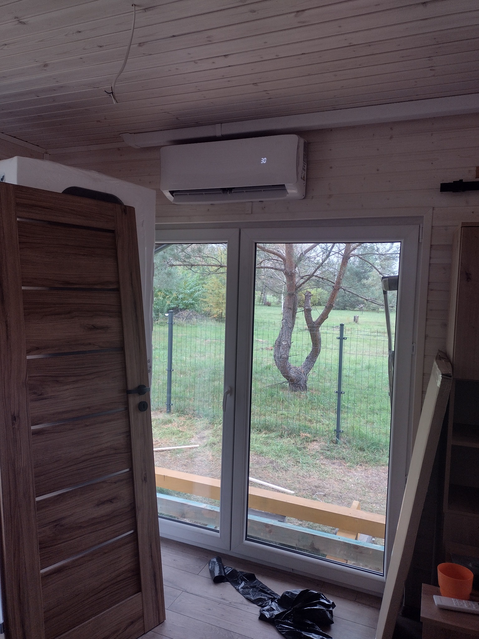 Biały klimatyzator ścienny zamontowany nad oknem w drewnianym domku, widok na ogród z drzewem przez okno, częściowo widoczna drewniana rama drzwi po lewej stronie.