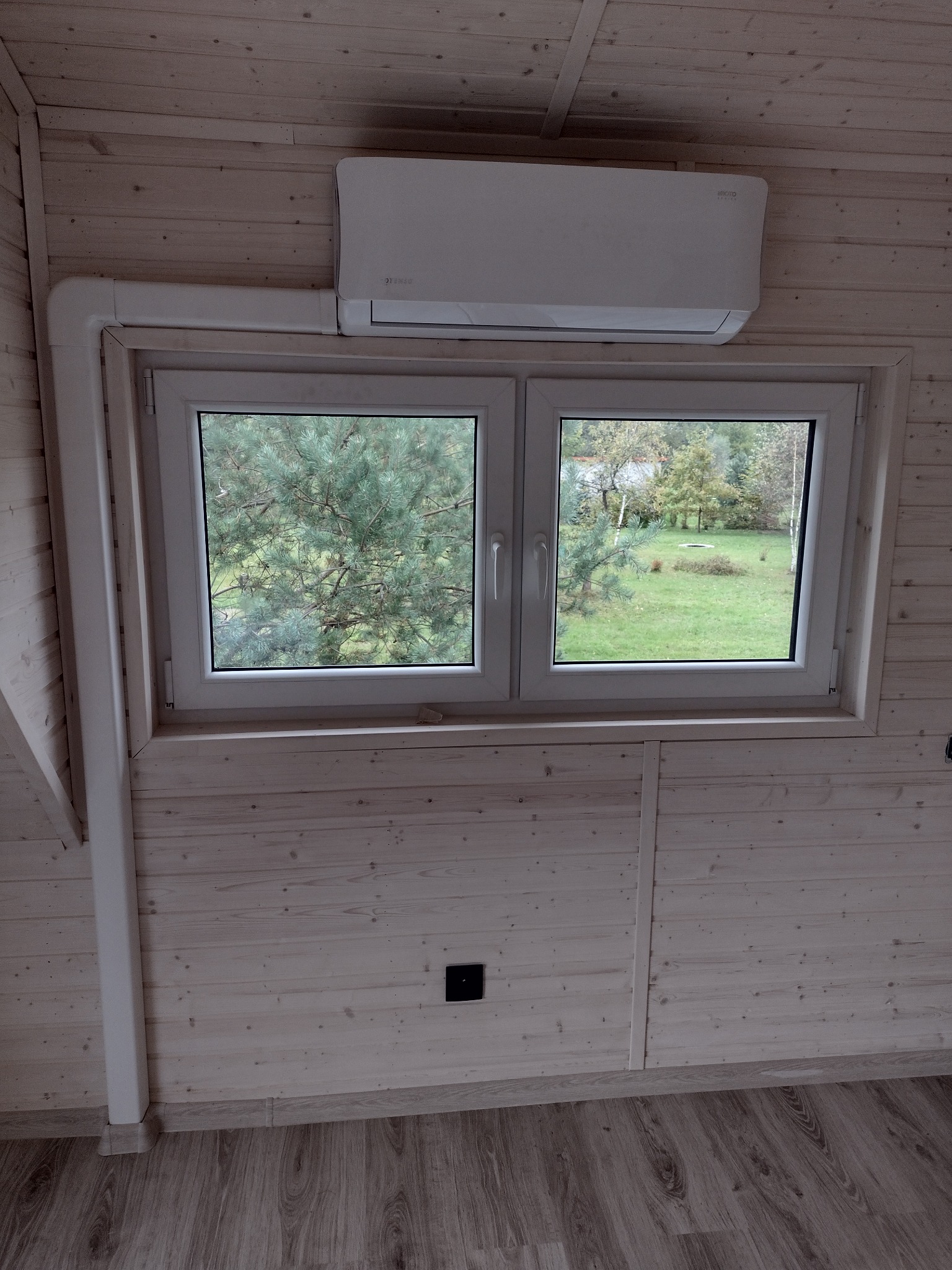 Biały klimatyzator zamontowany nad oknem w pokoju wykończonym jasnym drewnem, z widokiem na zielony ogród.