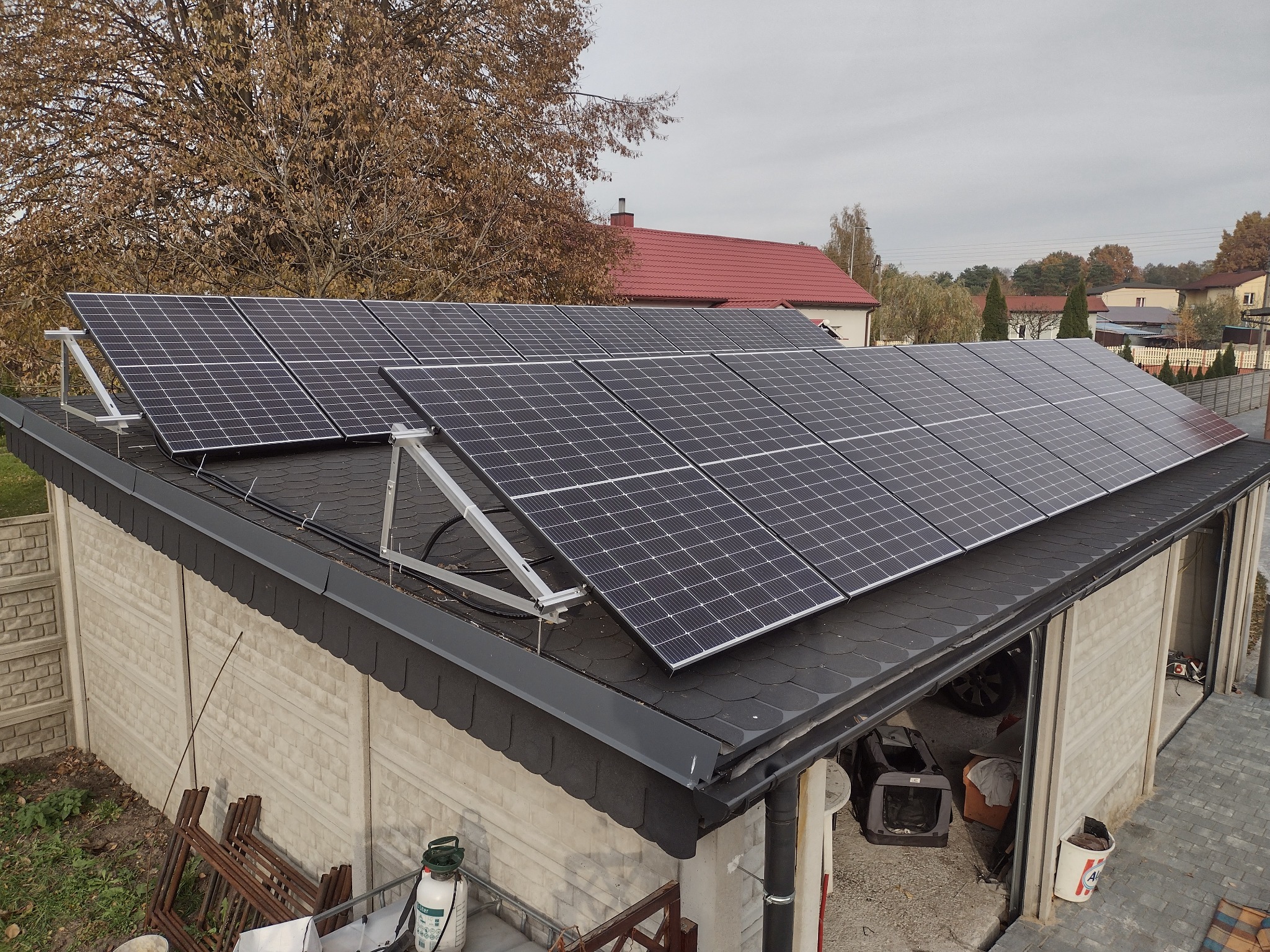 Panele słoneczne zamontowane na dachu garażu z bitumiczną dachówką, widoczne elementy konstrukcji mocującej, w tle drzewa jesienią i zabudowa jednorodzinna.