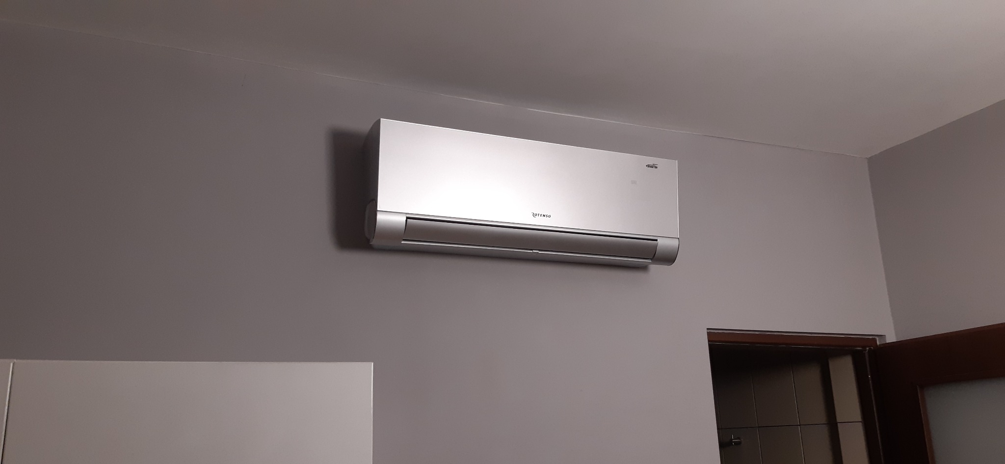 Nowoczesny, srebrny klimatyzator marki Rotenso, zamontowany na szarej ścianie w pomieszczeniu, widoczny fragment białej szafki i otworu drzwiowego.