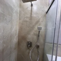 Beżowa kabina prysznicowa z deszczownicą i słuchawką, wykończona płytkami imitującymi marmur, z widocznym fragmentem wanny.