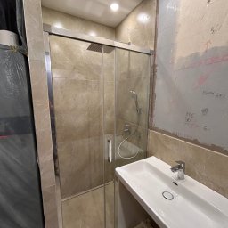 DDK PROF - Niedokończona łazienka z kabiną prysznicową z przesuwnymi drzwiami, umywalką i częściowo wykończonymi ścianami z widocznymi notatkami na ścianie.