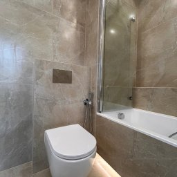 DDK PROF - Elegancka, wykończona łazienka w Świdnicy z beżowymi płytkami imitującymi marmur, podwieszana toaleta z chromowanym przyciskiem spłukiwania i szklanym parawanem nawannowym, oświetlenie LED pod wanną.