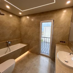 DDK PROF - Beżowa łazienka z podświetlaną wanną, umywalką i toaletą w zabudowie, z wyjściem na balkon, oświetlenie ledowe na suficie, widoczne przygotowane punkty elektryczne.