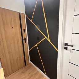 DDK PROF - Nowoczesny przedpokój z drewnianą podłogą, drzwi wejściowe z drewnopodobną okleiną i czarnymi akcentami, ściana pomalowana na czarno z geometrycznym wzorem z cienkich, złotych linii oraz białe...