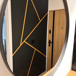 DDK PROF - Owalne lustro odbija fragment przedpokoju z czarną ścianą zdobioną geometrycznymi, złotymi liniami i drewnianymi drzwiami wejściowymi z czarnymi akcentami.