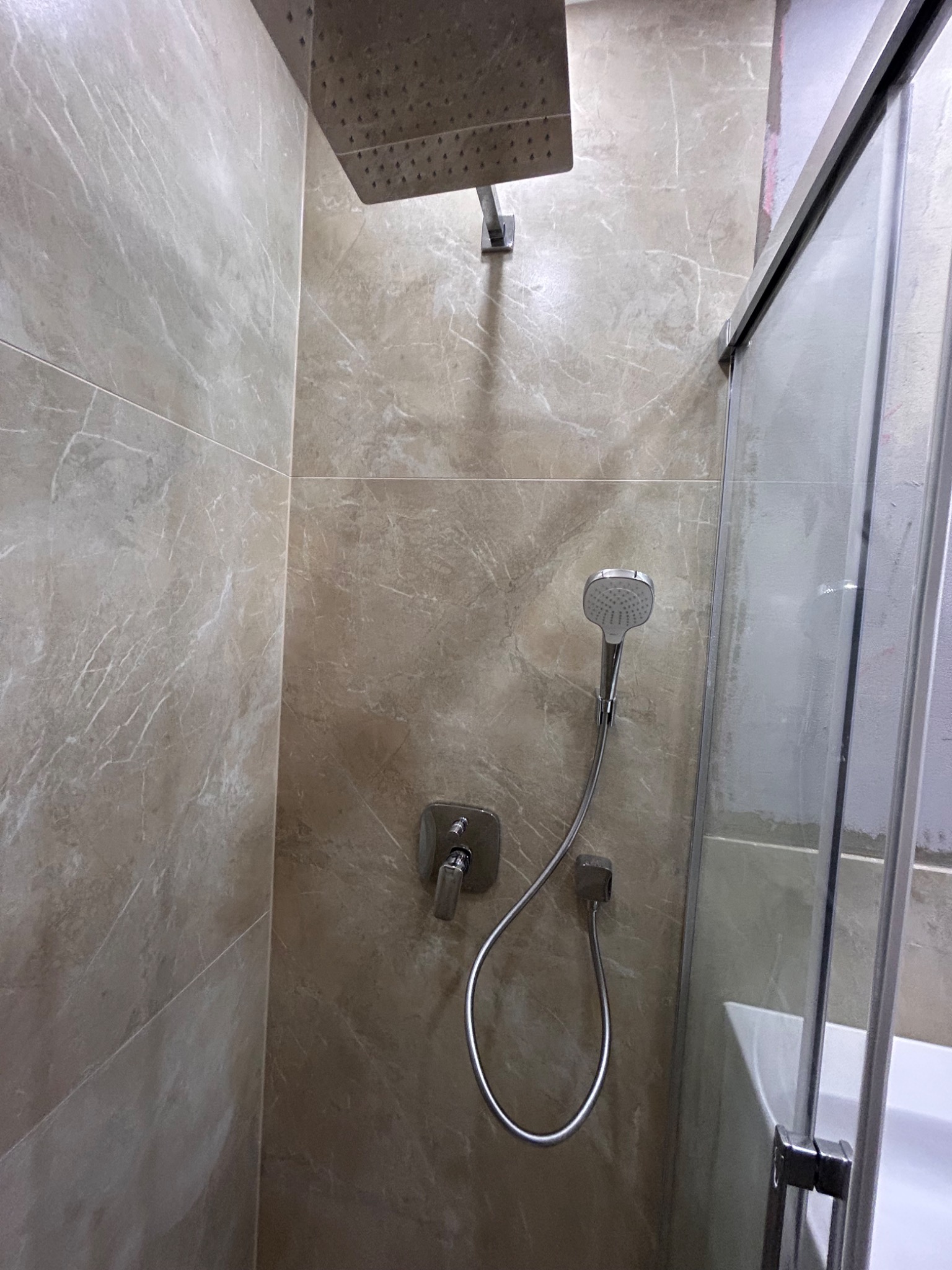 Beżowa kabina prysznicowa z deszczownicą i słuchawką, wykończona płytkami imitującymi marmur, z widocznym fragmentem wanny.