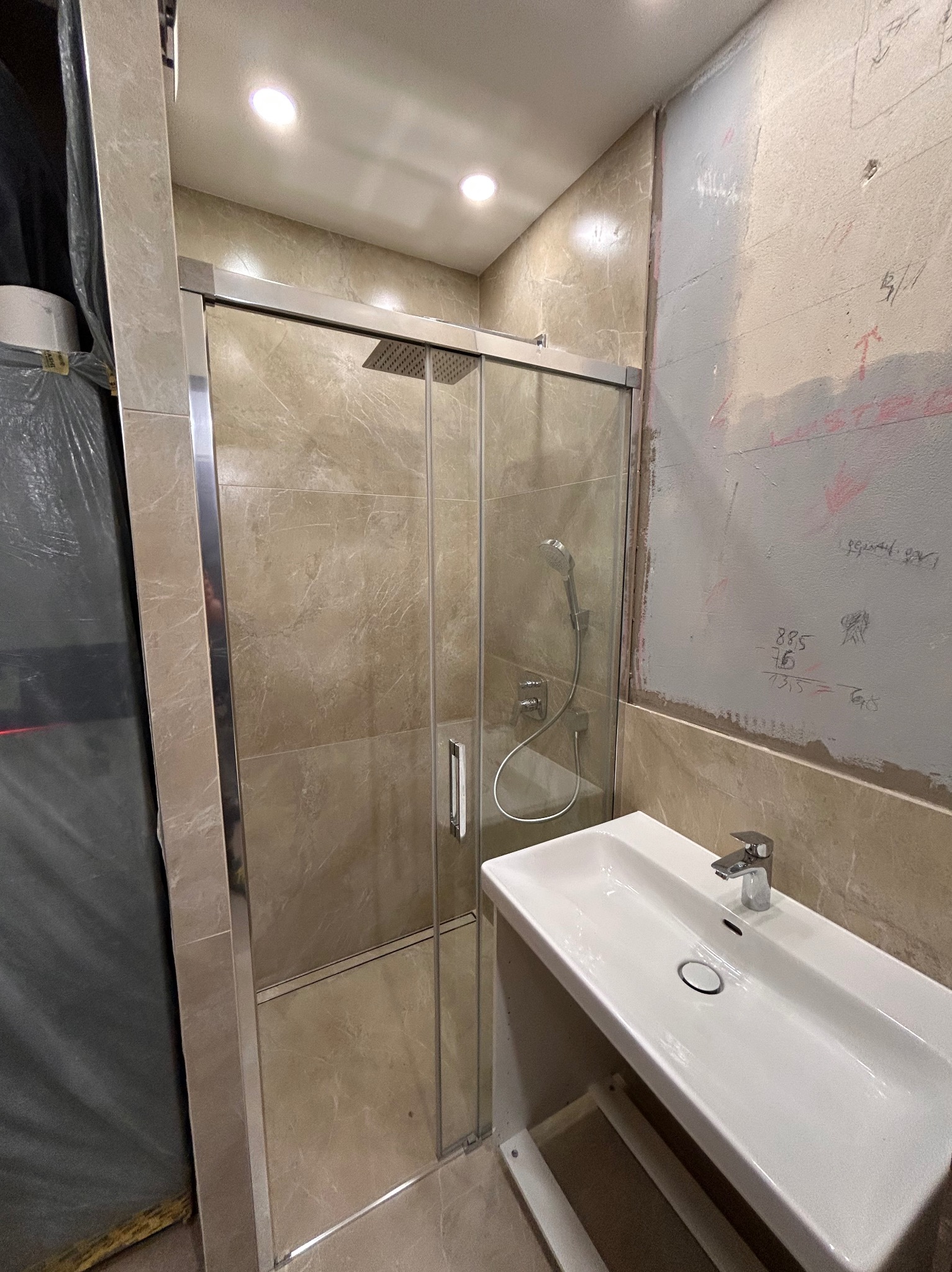 Niedokończona łazienka z kabiną prysznicową z przesuwnymi drzwiami, umywalką i częściowo wykończonymi ścianami z widocznymi notatkami na ścianie.