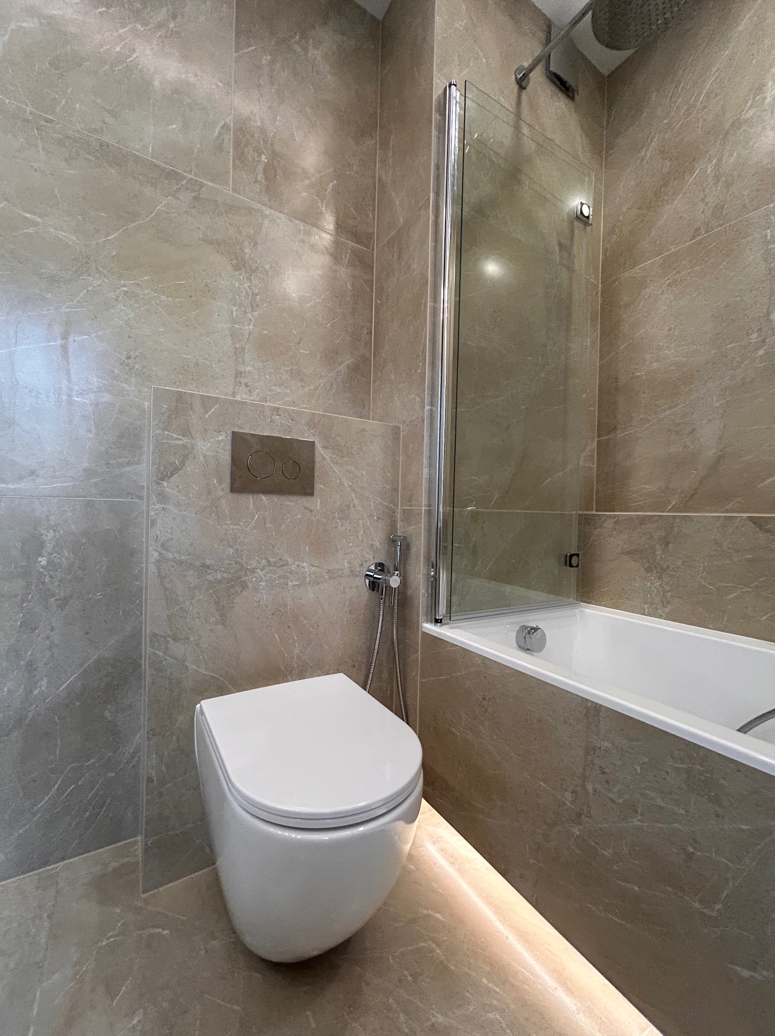 Elegancka, wykończona łazienka w Świdnicy z beżowymi płytkami imitującymi marmur, podwieszana toaleta z chromowanym przyciskiem spłukiwania i szklanym parawanem nawannowym, oświetlenie LED pod wanną.
