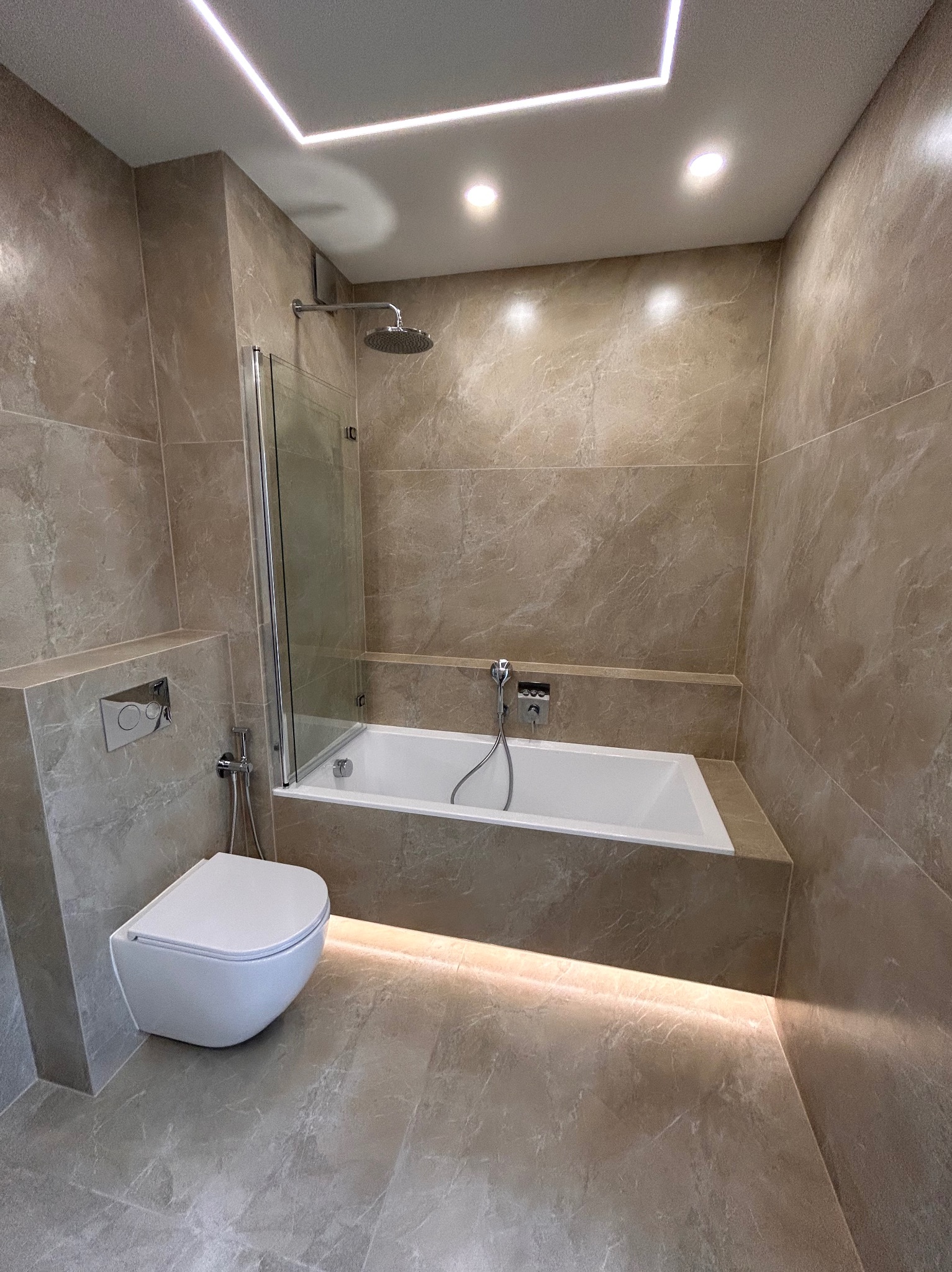 Beżowa łazienka z podświetleniem LED, wiszącą toaletą i wanną z parawanem nawannowym, wykończona marmurowymi płytkami.