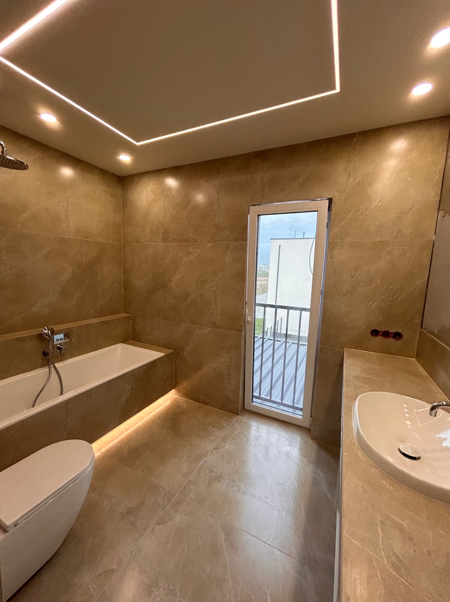 Beżowa łazienka z podświetlaną wanną, umywalką i toaletą w zabudowie, z wyjściem na balkon, oświetlenie ledowe na suficie, widoczne przygotowane punkty elektryczne.
