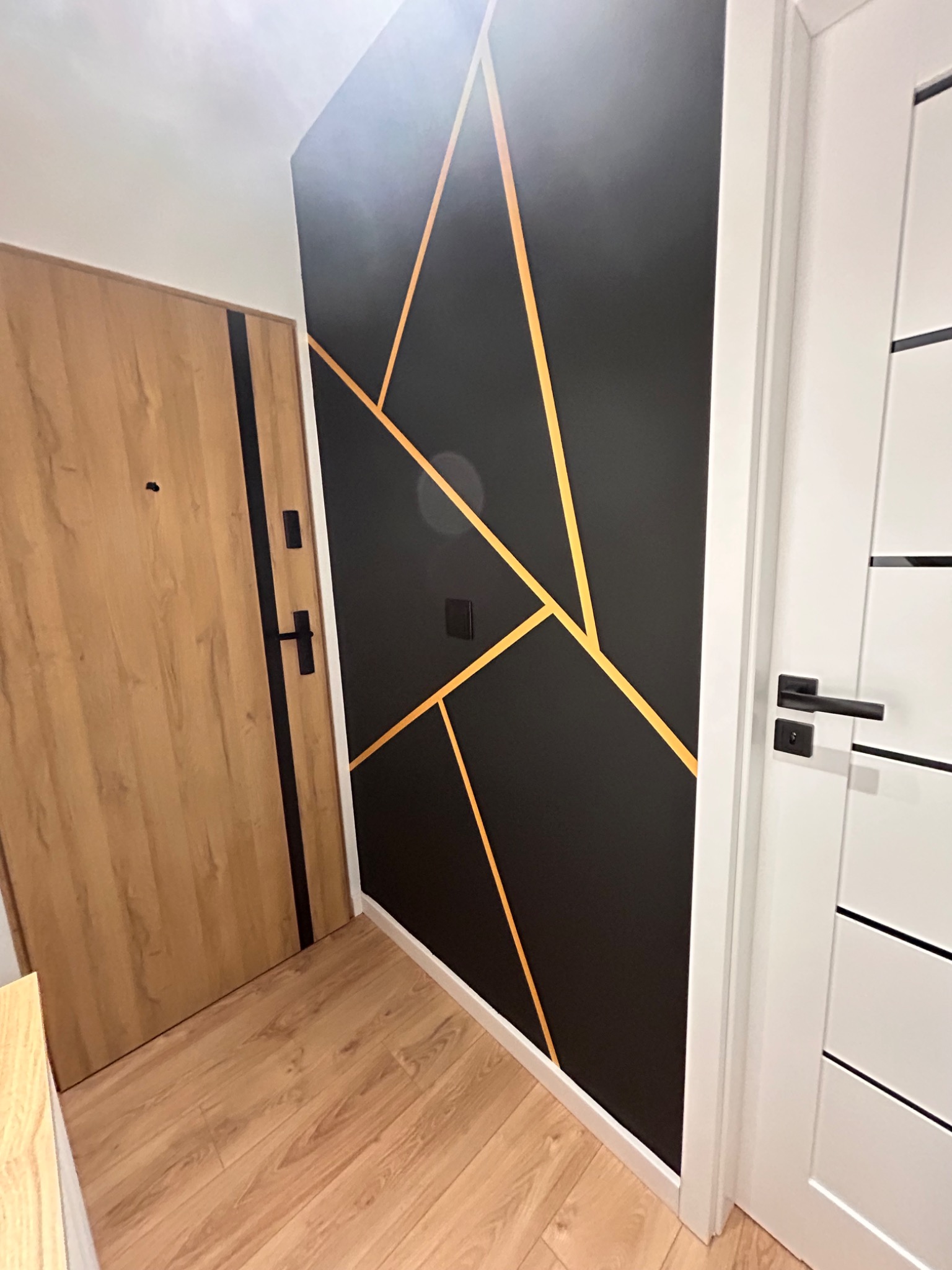 Nowoczesny przedpokój z drewnianą podłogą, drzwi wejściowe z drewnopodobną okleiną i czarnymi akcentami, ściana pomalowana na czarno z geometrycznym wzorem z cienkich, złotych linii oraz białe...