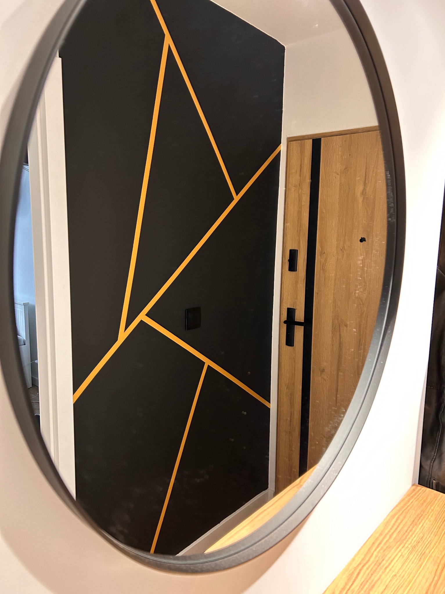 Owalne lustro odbija fragment przedpokoju z czarną ścianą zdobioną geometrycznymi, złotymi liniami i drewnianymi drzwiami wejściowymi z czarnymi akcentami.