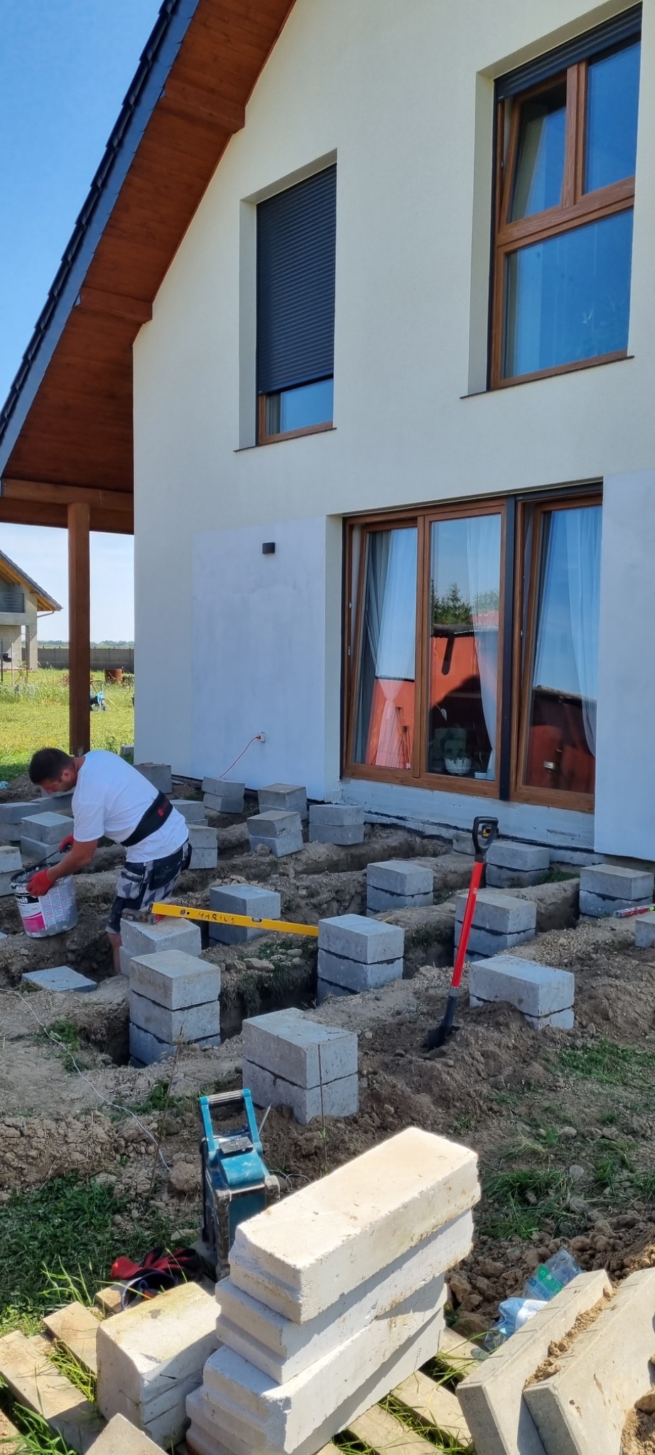 Pracownik w trakcie przygotowywania fundamentów pod dom szkieletowy, układanie betonowych bloczków na ziemi, widoczny poziomica i ubijak do gruntu.