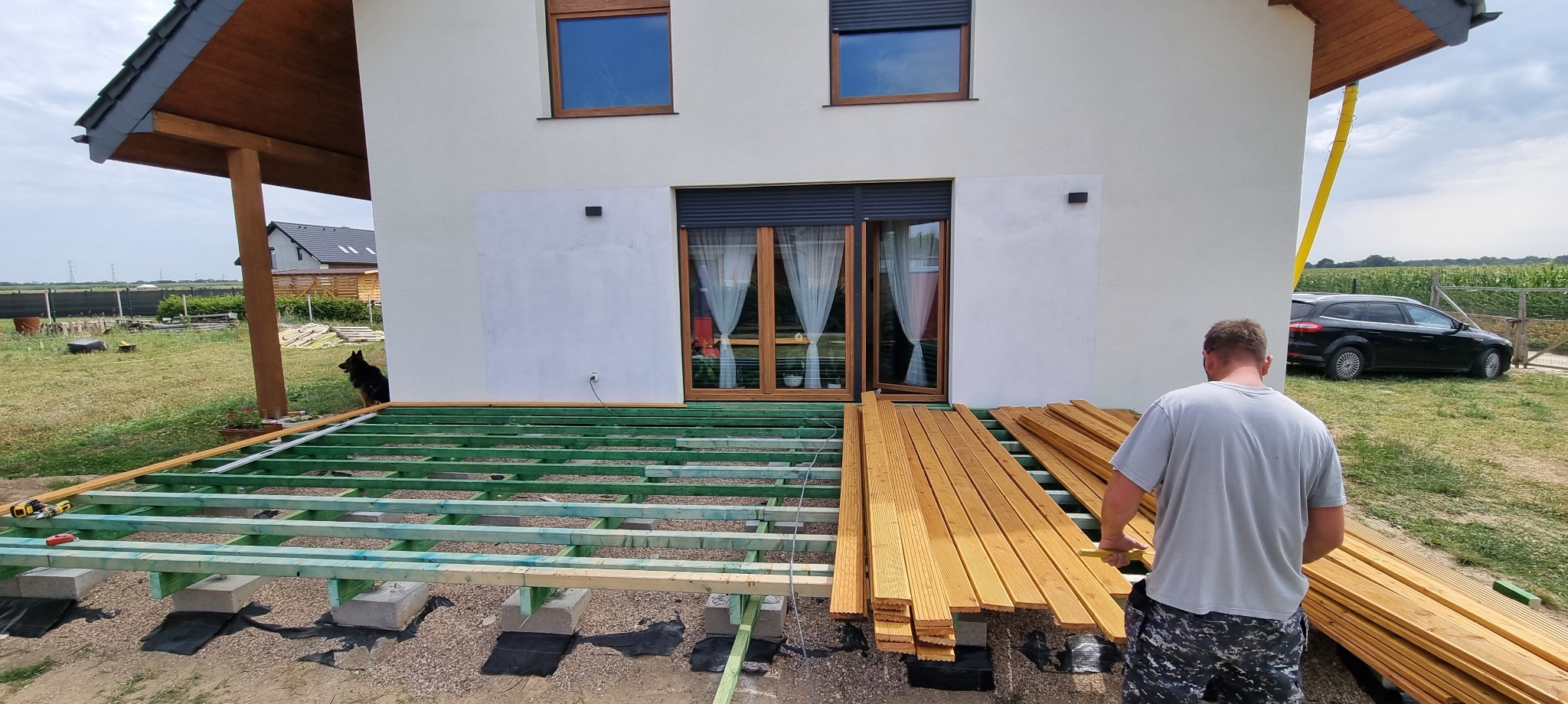 Budowa drewnianego tarasu przy białym domu jednorodzinnym: widoczny szkielet konstrukcji z impregnowanego drewna, stos desek tarasowych, pracownik z miarką, czarny pies obserwujący prace.