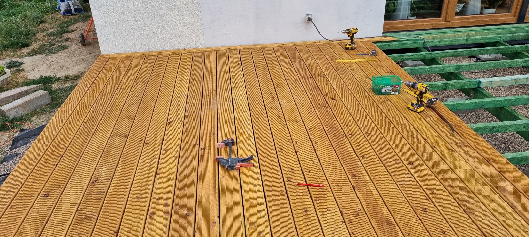 Drewniany taras w trakcie budowy, widoczne narzędzia: wkrętarka, ściski stolarskie, ołówek, poziomica, puszka wkrętów, zielona konstrukcja nośna, fragment elewacji budynku z gniazdkiem elektrycznym.