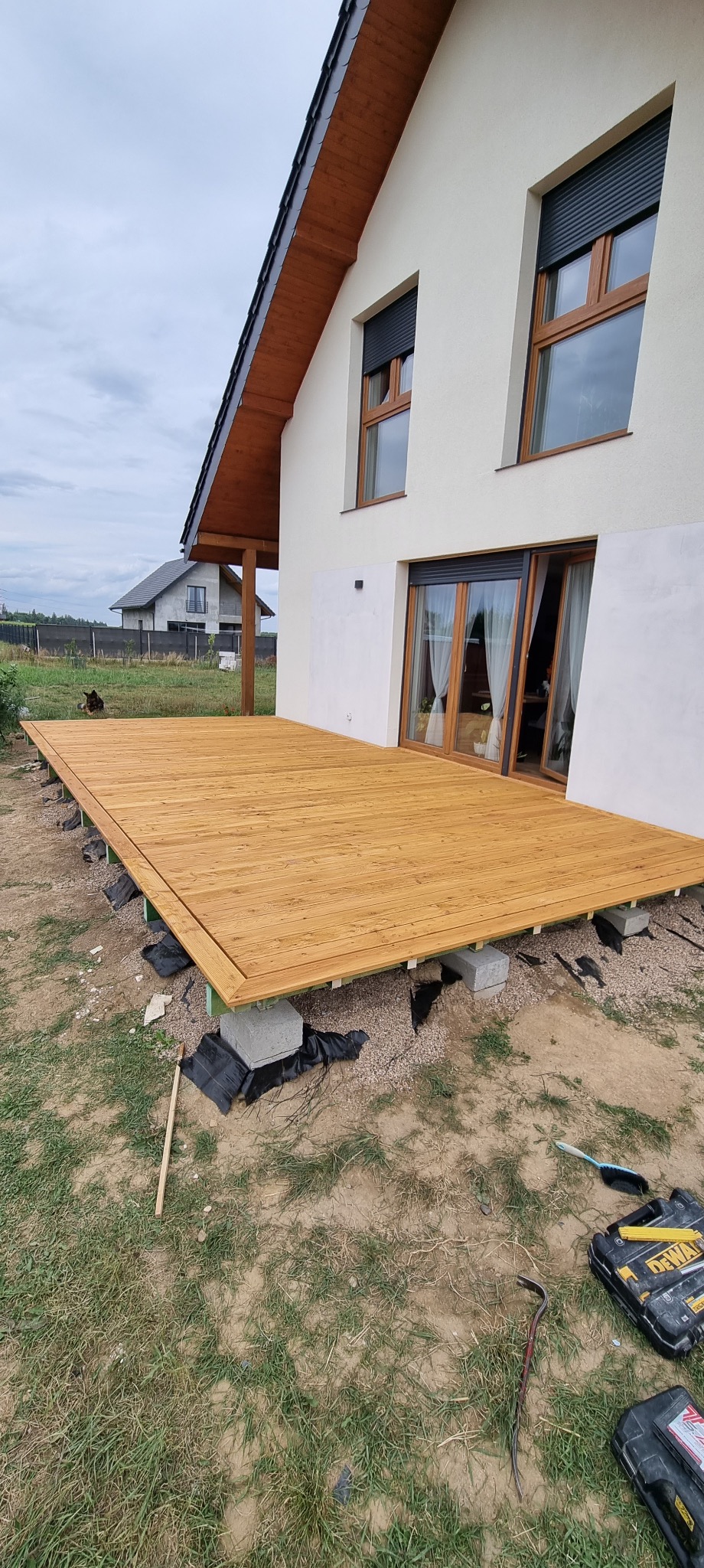 Drewniany taras w trakcie budowy przy nowoczesnym domu z białymi ścianami, widoczne bloczki betonowe pod konstrukcją i narzędzia na ziemi.