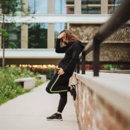 Mężczyzna w czarnym dresie z kapturem i żółto-niebieskim paskiem na spodniach, opierający się o mur na tle nowoczesnego budynku i zieleni.