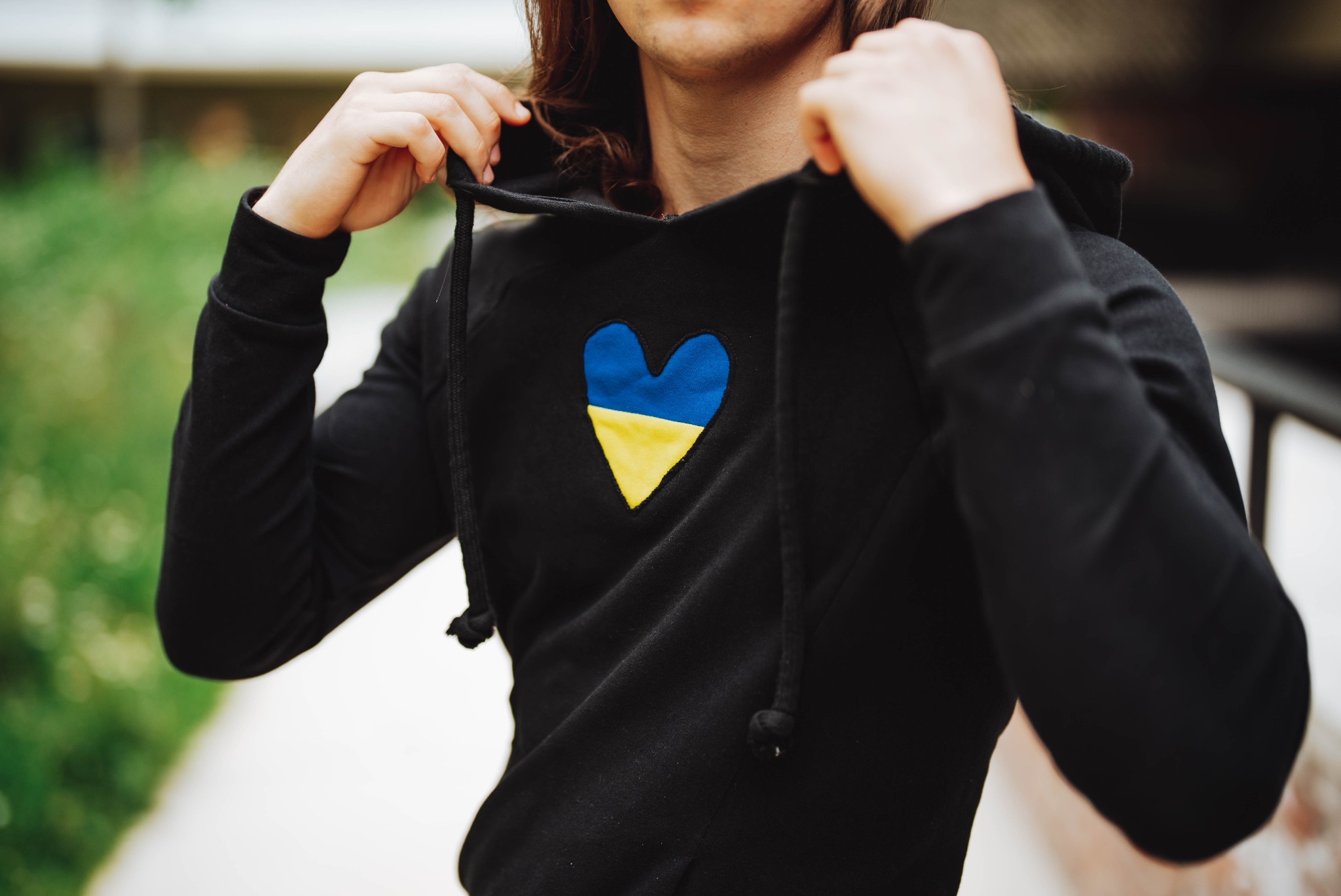 Czarna bluza z kapturem, z naszytym sercem w barwach flagi Ukrainy, prezentowana na osobie z długimi włosami, na tle zieleni.