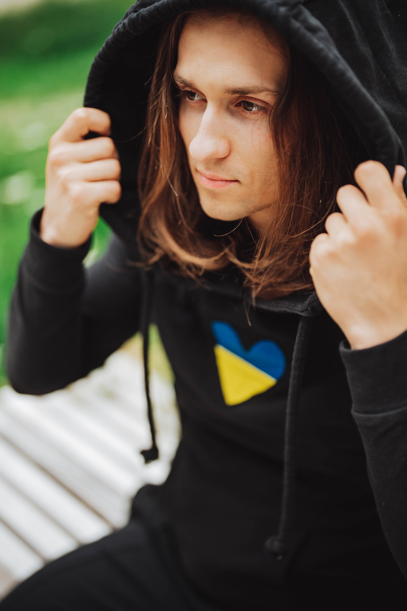 Młody mężczyzna z długimi włosami w czarnej bluzie z kapturem i naszytym sercem w barwach flagi Ukrainy, siedzący na ławce.