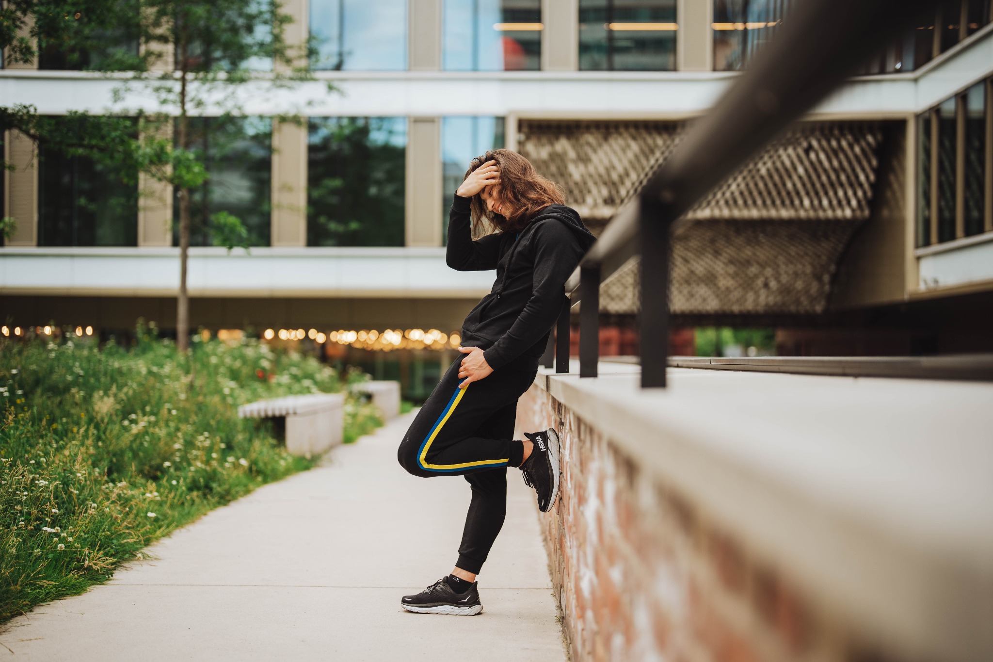 Mężczyzna w czarnym dresie z kapturem i żółto-niebieskim paskiem na spodniach, opierający się o mur na tle nowoczesnego budynku i zieleni.