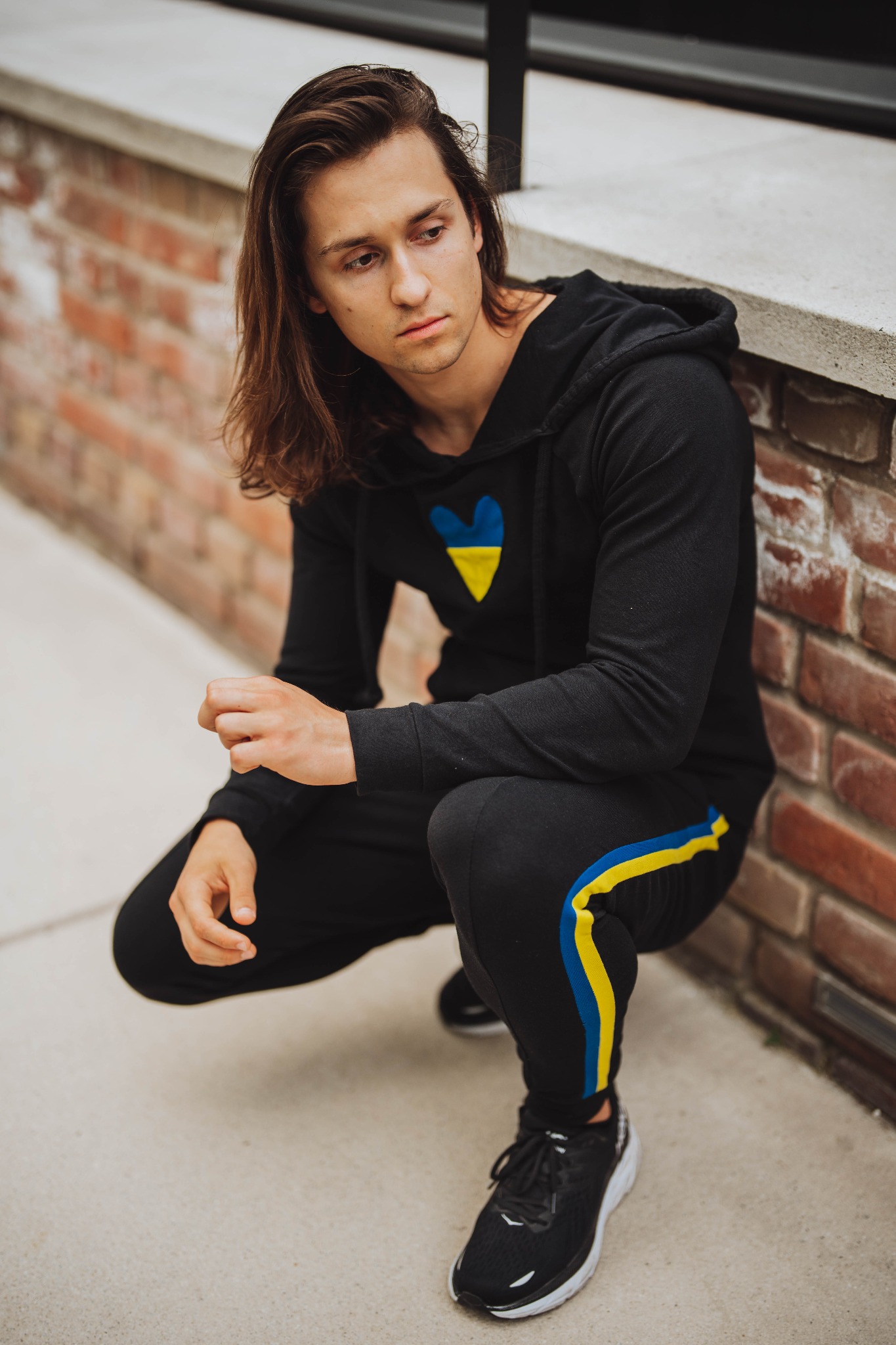 Mężczyzna w czarnym dresie z kapturem i ukraińskim motywem serca, kuca przy ceglanej ścianie, spoglądając w dół.