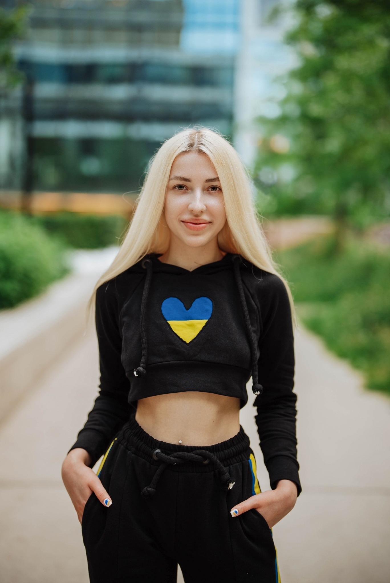 Blondwłosa młoda kobieta w czarnym, krótkim hoodie z naszytym sercem w barwach flagi Ukrainy i czarnych spodniach dresowych z żółtymi lampasami, pozująca na tle miejskiego parku.