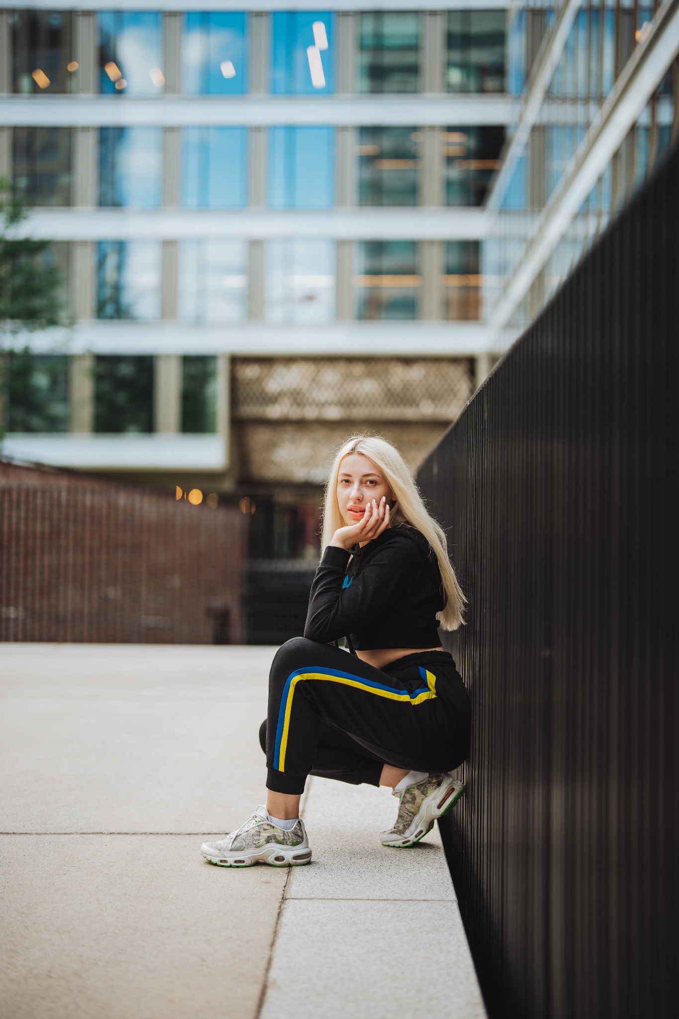 Młoda kobieta z blond włosami w czarnym dresie z żółto-niebieskimi paskami, pozująca w kucki na tle nowoczesnego budynku z przeszkleniami.